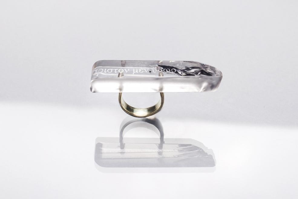 Transparent rectangular ring with embedded text and a dark decorative element, placed on a light background with reflection.