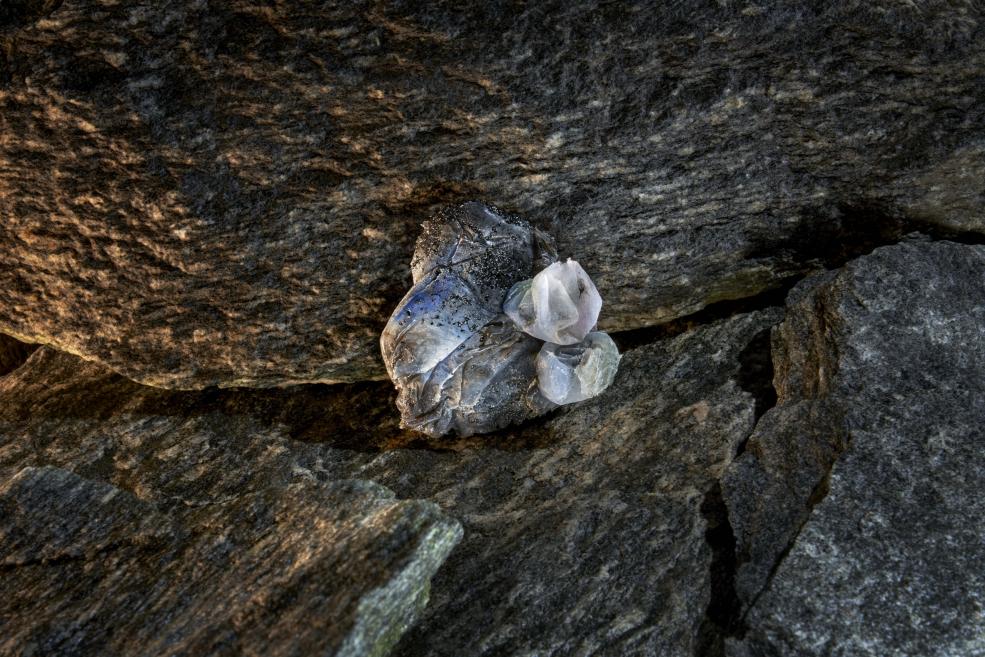 A transparent piece of plastic debris with sand grains trapped between rocks in a natural setting.