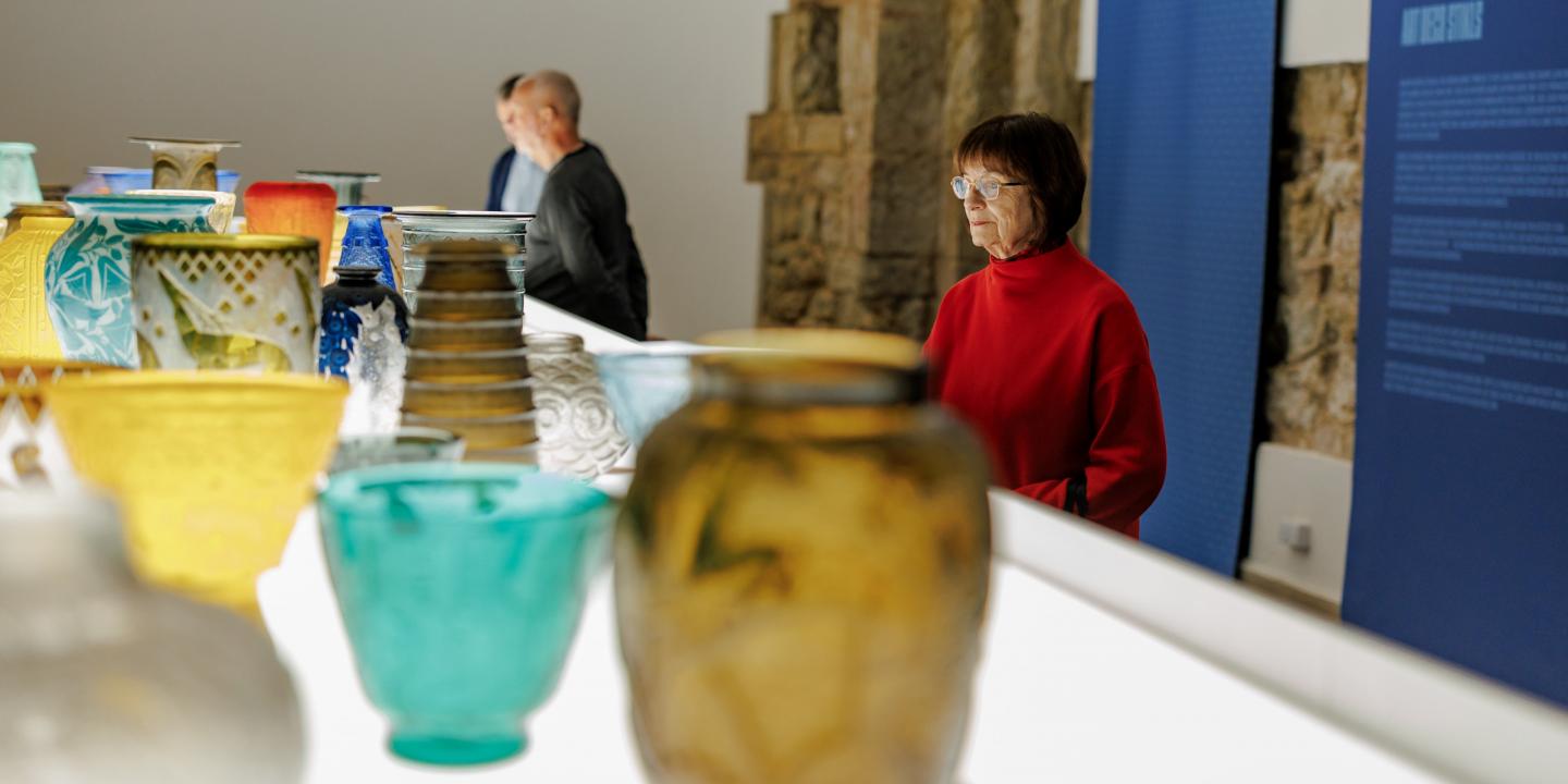 Muzeja ekspozīcijā izstādīti krāsaini stikla trauki, kamēr apmeklētāji tos aplūko telpā ar informatīvu stendu.