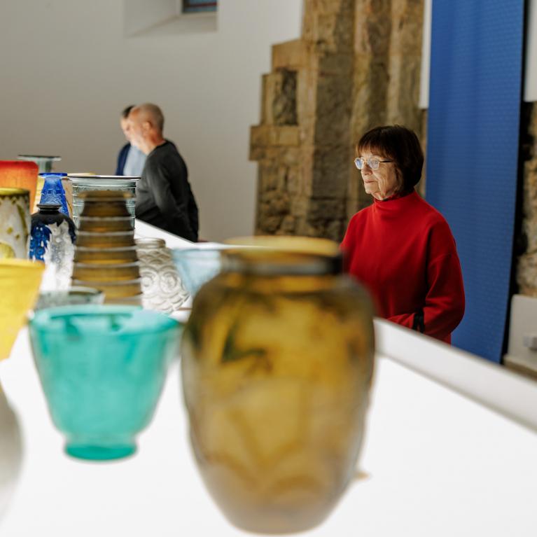 Muzeja ekspozīcijā izstādīti krāsaini stikla trauki, kamēr apmeklētāji tos aplūko telpā ar informatīvu stendu.