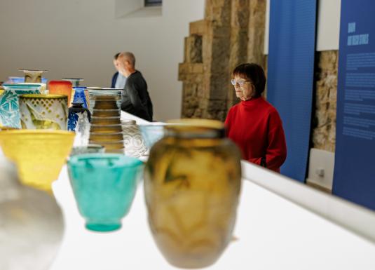 Muzeja ekspozīcijā izstādīti krāsaini stikla trauki, kamēr apmeklētāji tos aplūko telpā ar informatīvu stendu.