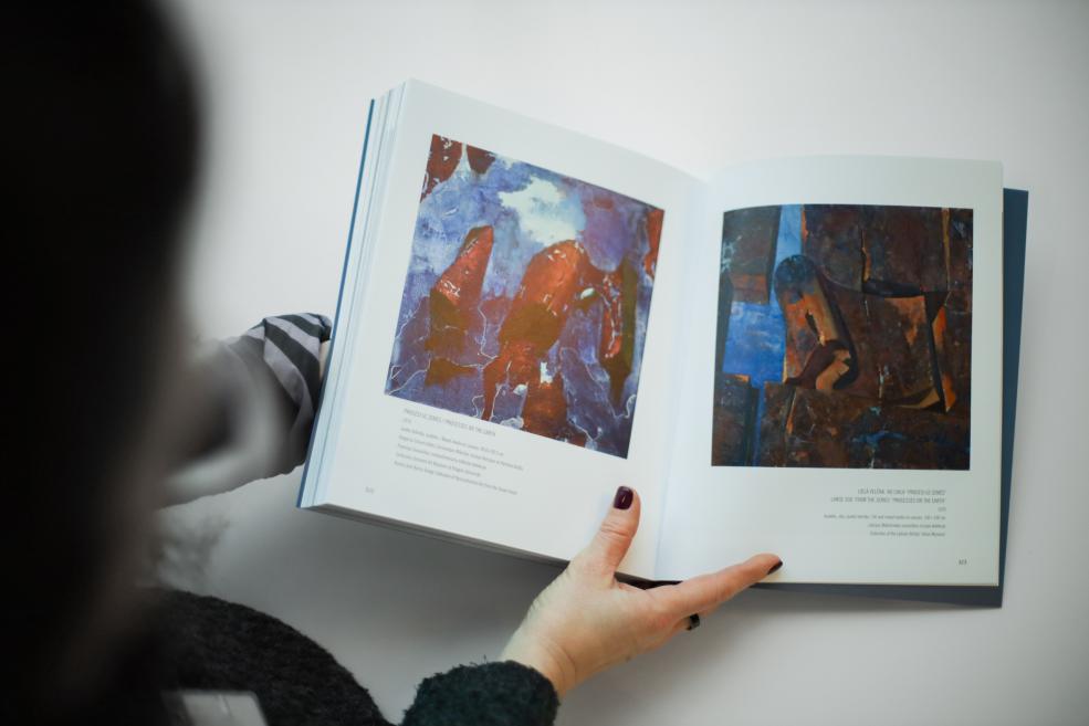 Persona tur atvērtu mākslas grāmatu, kurā redzami divi abstrakti gleznojumi zilgani sarkanā un tumšā krāsu gammā.