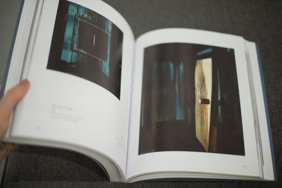 Grāmata &ldquo;Ojārs Ābols. Cilvēka absurdie projekti uz Zemes&rdquo; uz gleznotājam veltītās izstādes fona Latvijas Nacionālajā mākslas muzejā. 2026. Foto: Estere Rožkalne