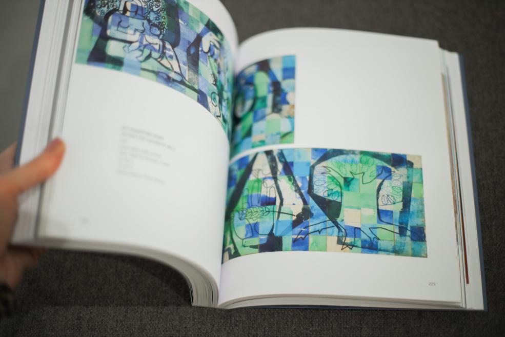 Persona pāršķir mākslas grāmatu, kur redzamas abstraktas kompozīcijas zilzaļos toņos ar ģeometriskiem un figurāliem elementiem.