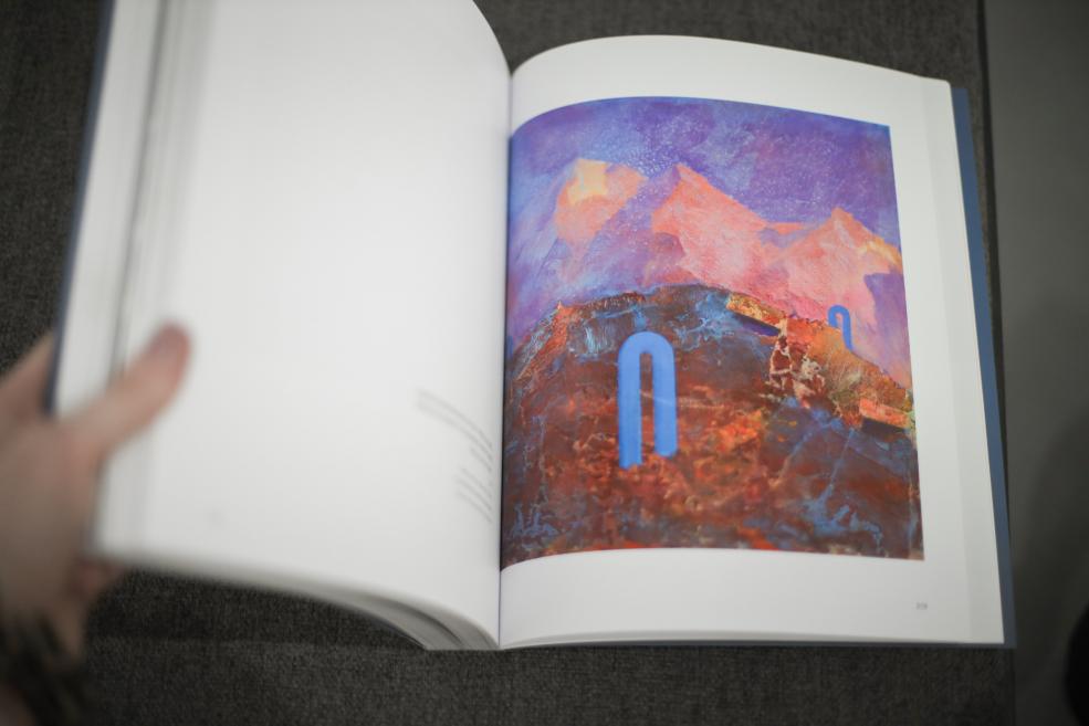 Persona pāršķir mākslas grāmatu, atklājot abstraktu gleznu ar kalnainu formu sarkanbrūnos un violetos toņos un zilu grafisku zīmi centrā.