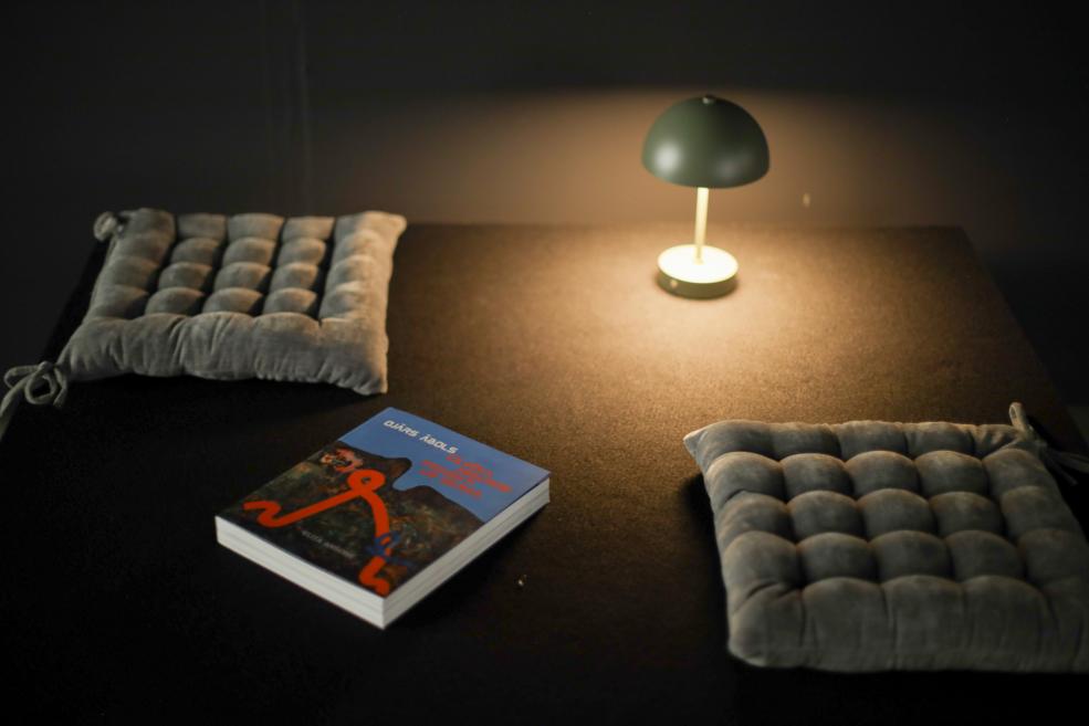 Tumša lasītavas telpa ar galdu, uz kura atrodas ieslēgta galda lampa, divi pelēki sēdēšanas spilveni un mākslas grāmata ar Ojāra Ābola darbu uz vāka.