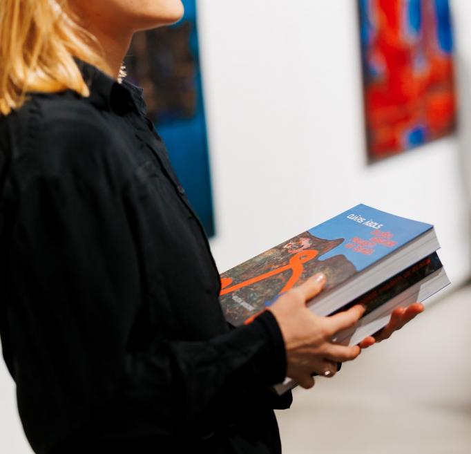 Sieviete mākslas izstāžu telpā rokās tur mākslas grāmatu ar Ojāra Ābola darbu uz vāka, fonā redzami gleznas.