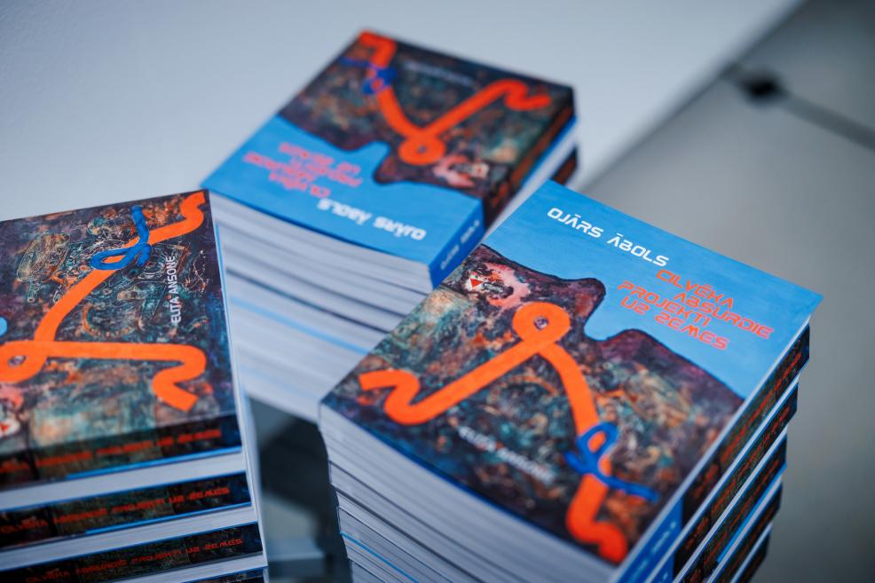 Uz galda sakrautas vairākas mākslas grāmatas “Ojārs Ābols. Cilvēka absurdie projekti uz zemes” ar zilām vākiem un oranžu grafisku motīvu.