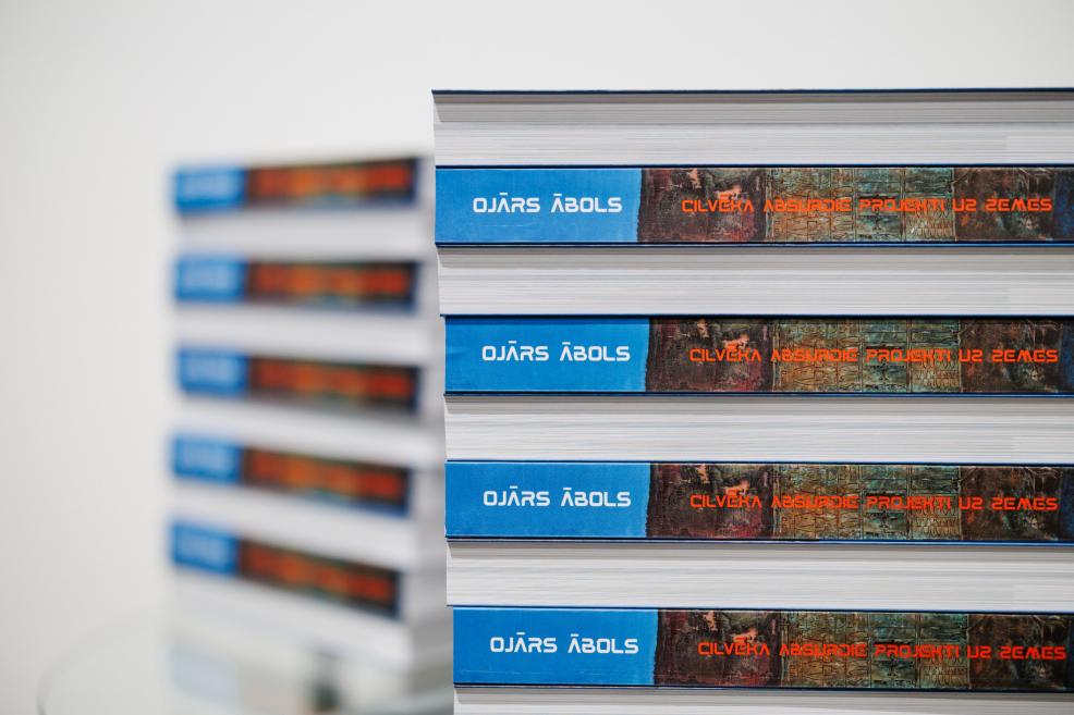 Kaudzē sakrautas mākslas grāmatas “Ojārs Ābols. Cilvēka absurdie projekti uz zemes”, redzami zilie muguriņu nosaukumi ar oranžu tipogrāfiju.