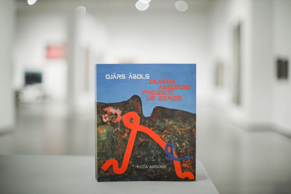 Uz pjedestāla izstādīta grāmata “Ojārs Ābols. Cilvēka absurdie projekti uz zemes” ar abstraktu, tumšu ainavu un spilgtu oranži zilu līniju uz zilgana fona, fonā redzama izplūdusi izstāžu telpa.