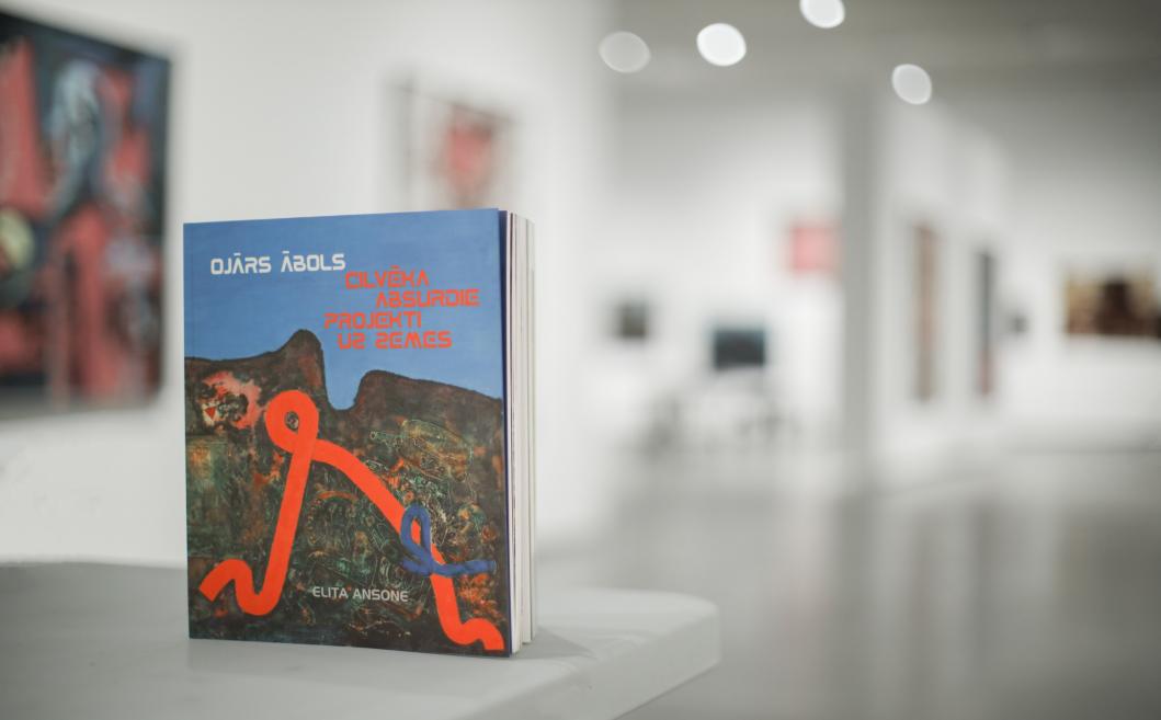 Uz pjedestāla izstādīta grāmata “Ojārs Ābols. Cilvēka absurdie projekti uz zemes” ar abstraktu, tumšu ainavu un spilgtu oranži zilu līniju uz zilgana fona, fonā redzama izplūdusi izstāžu telpa.