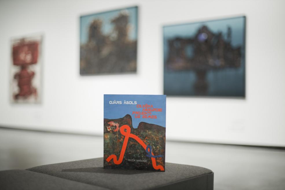 Uz pjedestāla izstādīta grāmata “Ojārs Ābols. Cilvēka absurdie projekti uz zemes” ar abstraktu, tumšu ainavu un spilgtu oranži zilu līniju uz zilgana fona, fonā redzama izplūdusi izstāžu telpa.