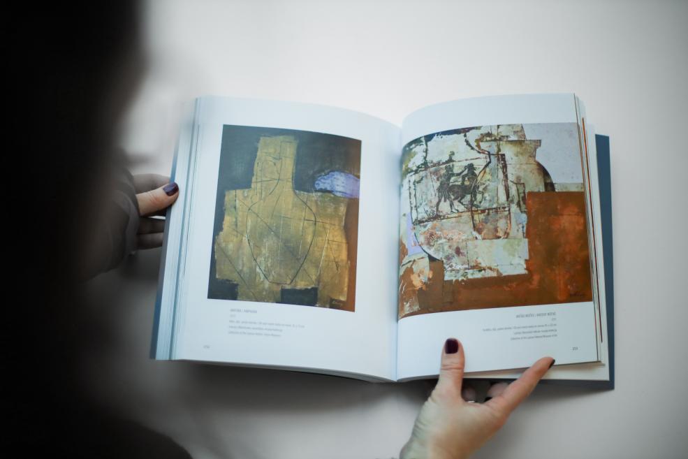 Persona tur rokās atvērtu mākslas grāmatu, kuras atvērumā redzamas divas abstraktas gleznas ar zemes toņu kompozīcijām uz gaiša fona.