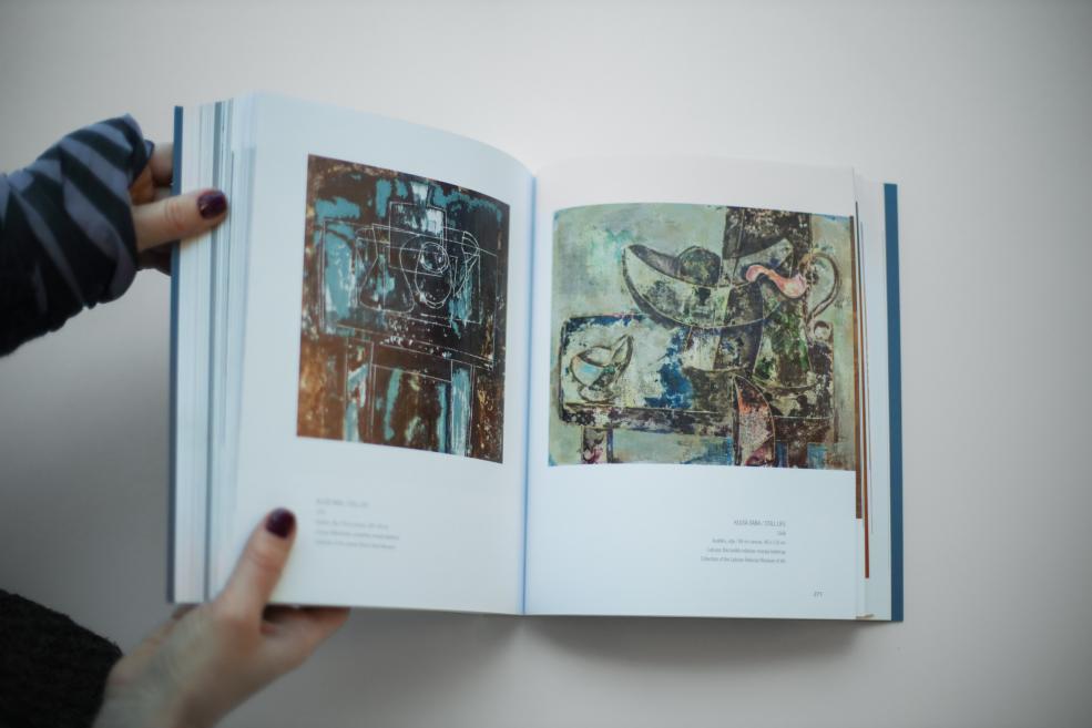 Persona tur atvērtu mākslas grāmatu, kuras atvērumā redzami divi abstrakti darbi ar daudzslāņainām formām un kontrastējošiem zilzaļiem un zemes toņiem.