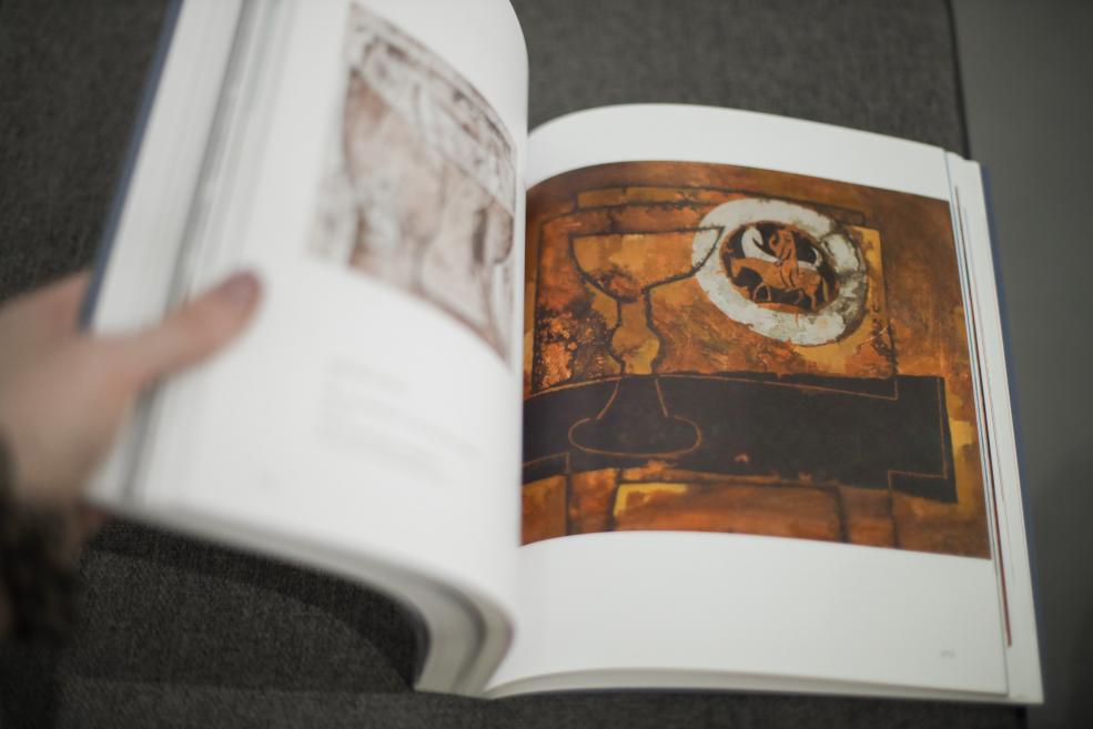 Grāmata &ldquo;Ojārs Ābols. Cilvēka absurdie projekti uz Zemes&rdquo; uz gleznotājam veltītās izstādes fona Latvijas Nacionālajā mākslas muzejā. 2026. Foto: Estere Rožkalne