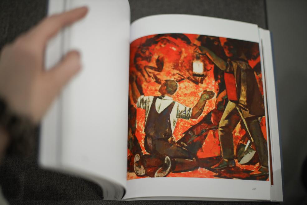 Persona šķirsta mākslas grāmatu, un labajā lapā redzama figurāla kompozīcija sarkanā krāsu gammā ar divām cilvēka figūrām, no kurām viena ir nometusies ceļos.