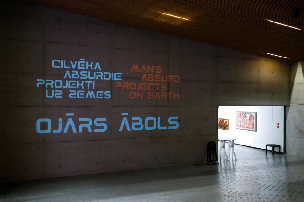An exhibition wall with the projected title “Human Absurd Projects on Earth. Ojārs Ābols,” with an entrance to the gallery space and paintings visible on the right.