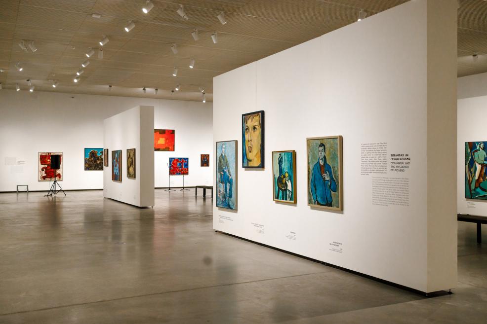 View of an exhibition gallery with white walls displaying paintings by Ojārs Ābols, accompanied by wall texts and benches in a spacious, open exhibition space.