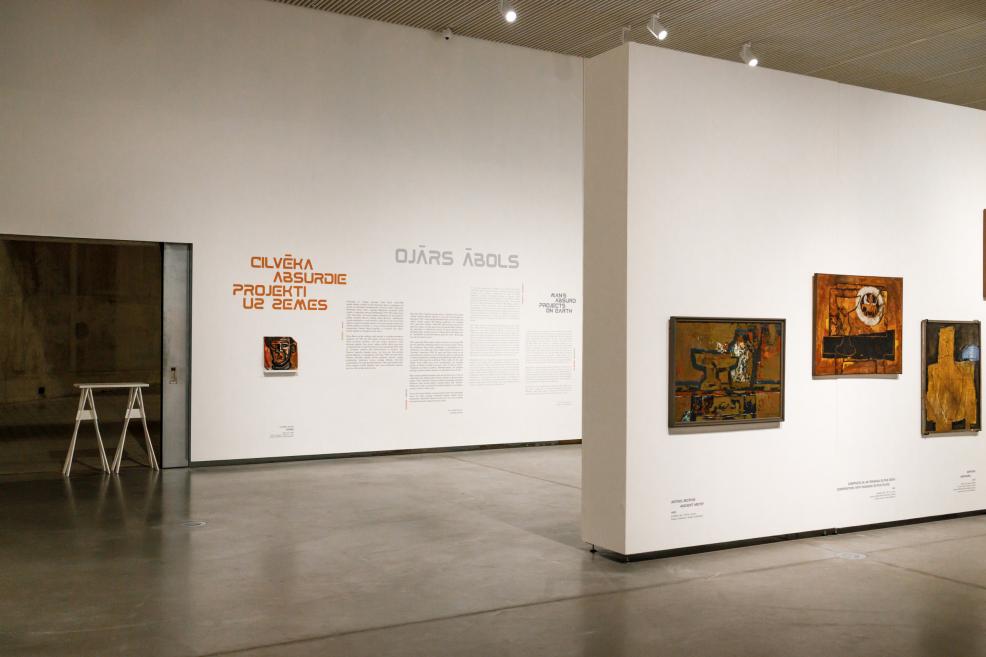 Exhibition view featuring wall texts and paintings by Ojārs Ābols displayed on white partition walls in a spacious, light-filled museum gallery.