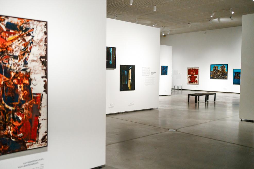 View of the exhibition gallery with paintings by Ojārs Ābols displayed on white partition walls, showing works in various formats, a bench for visitors, and an open, well-lit space.