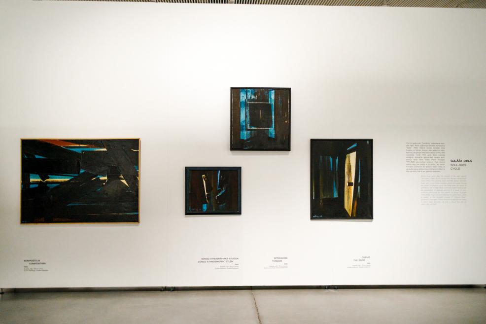 Detail of an exhibition wall featuring four dark-toned paintings by Ojārs Ābols displayed on a white wall, with accompanying exhibition text visible nearby.