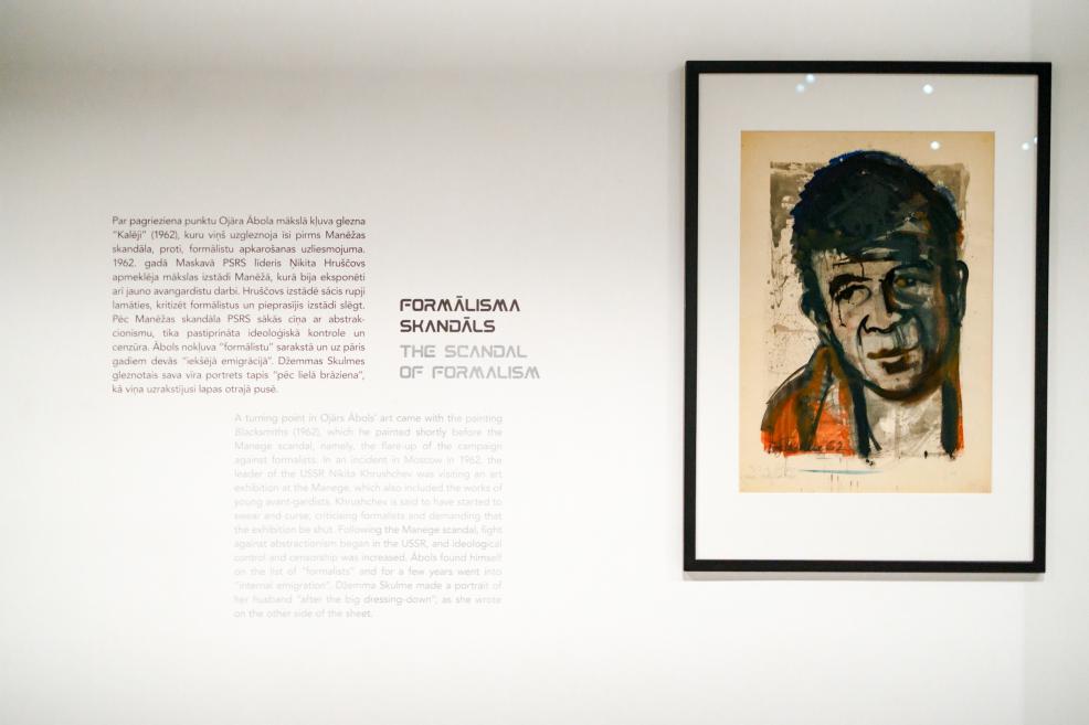 Izstādes siena ar informatīvu tekstu par formālisma skandālu un ierāmētu Ojāra Ābola portreta gleznu uz baltas sienas.