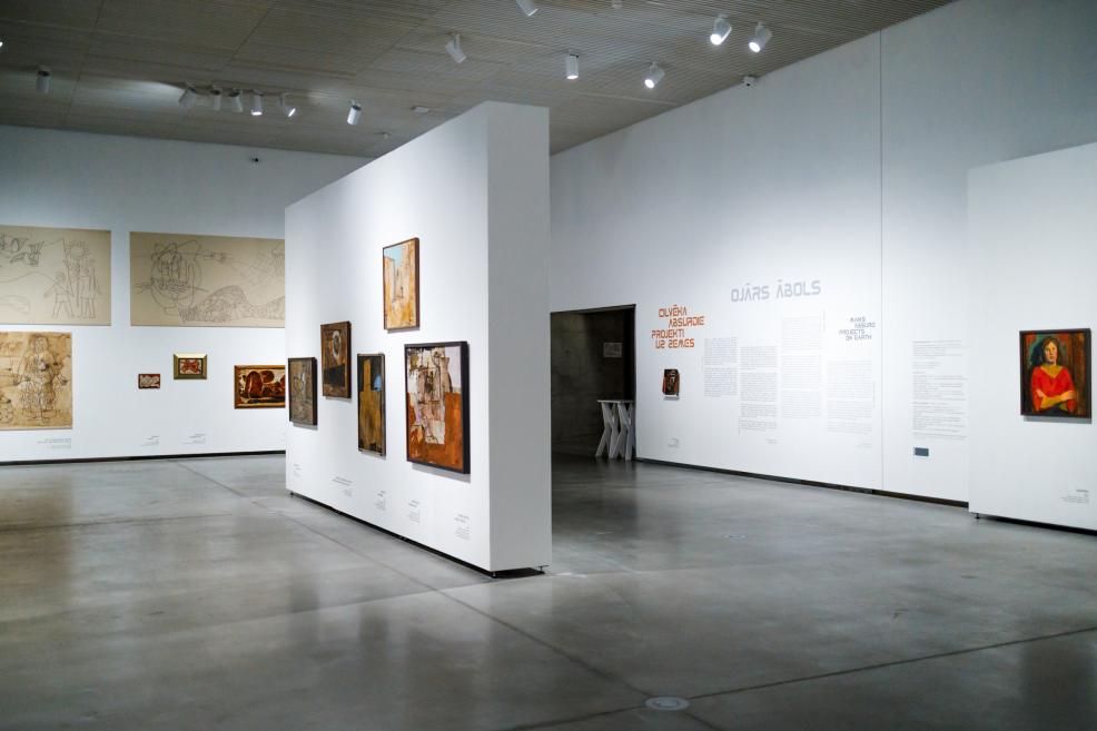 Exhibition overview showing works by Ojārs Ābols displayed on white walls with informational texts and drawings in a spacious museum gallery with overhead lighting.