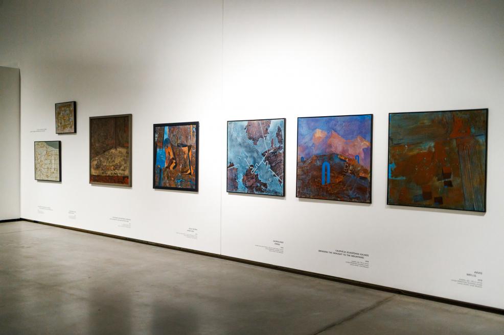 Section of an exhibition wall featuring several framed paintings by Ojārs Ābols arranged in a row on a white wall, with artwork titles and descriptions displayed below.