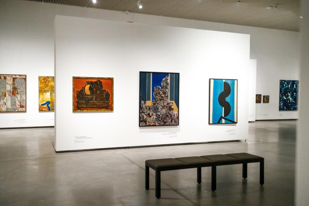 Exhibition space with a white wall displaying several framed paintings by Ojārs Ābols; a black bench is visible in the foreground, with a spacious museum gallery and additional artworks in the background.