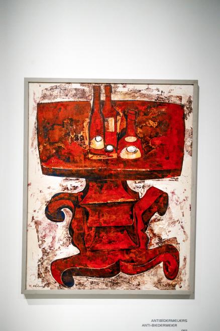 Painting by Ojārs Ābols depicting a stylized red table with bottles and vessels, executed in an expressive, textured manner against a contrasting white background.