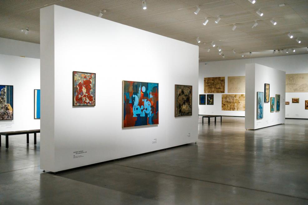 View of an exhibition gallery with white partition walls displaying paintings by Ojārs Ābols, featuring high ceilings, directional lighting, and benches placed throughout the open space.