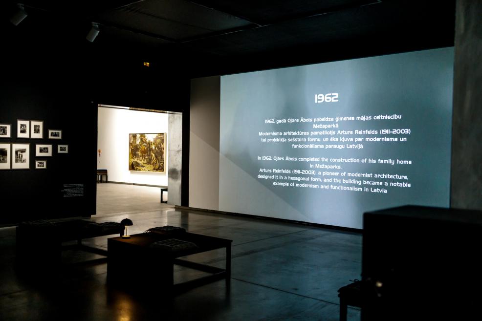 Dimly lit exhibition space with wall-projected text “1962” describing a key moment in Ojārs Ābols’s life, benches in the foreground, and a lit painting visible in an adjacent gallery area.