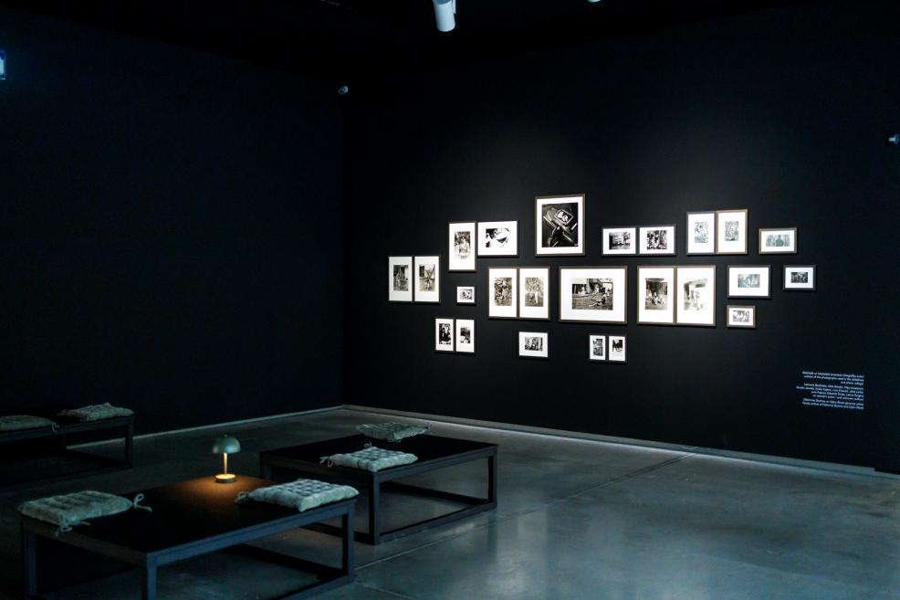 Dark exhibition room with black walls displaying a grouped arrangement of black-and-white photographs in various sizes; low tables with cushions and a small lamp in the center create a quiet, contemplative atmosphere.