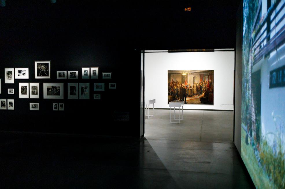 Tumša izstādes telpa ar melnām sienām; kreisajā pusē – melnbaltu fotogrāfiju kompozīcija, bet caur atvērumu redzama izgaismota blakustelpa ar monumentālu vēsturisku gleznu un diviem informatīviem stendiem, radot kontrastu starp tumsu un gaismu.