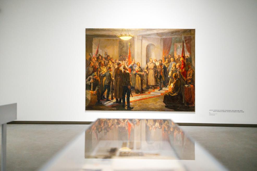 Izstāžu zālē uz baltas sienas eksponēta monumentāla vēsturiska glezna ar cilvēku grupu karogiem rotātā telpā; priekšplānā redzams stikla vitrīnas atspīdums, kas pastiprina kompozīcijas simetriju un gaismu.