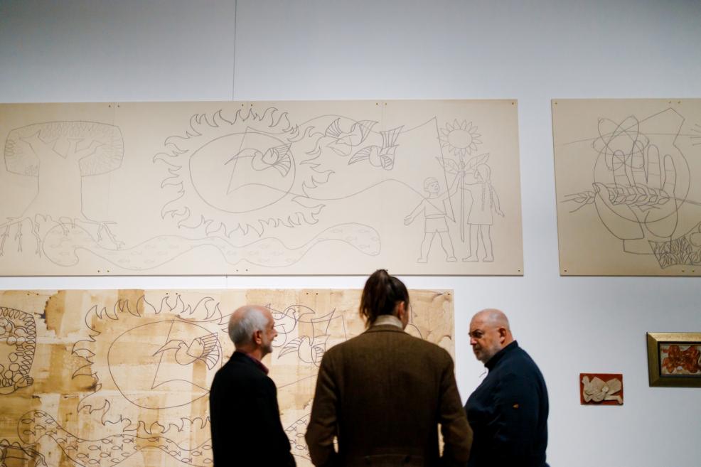 Trīs apmeklētāji stāv izstāžu zālē un aplūko uz sienas eksponētus lielizmēra grafiskus zīmējumus ar simboliskiem motīviem – sauli, putniem, augiem un bērnu figūrām.
