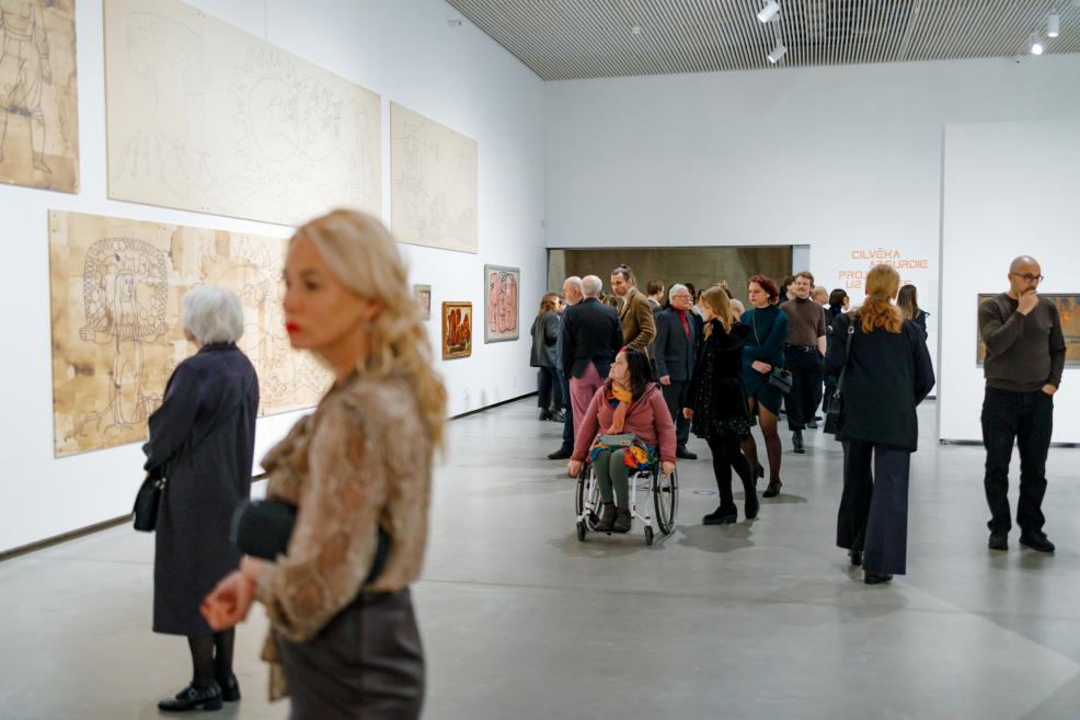 Several visitors move through a gallery space viewing artworks on the walls; a blurred female figure in the foreground and, in the background, a group of people—including a wheelchair user—gathered around large-scale graphic works and paintings.