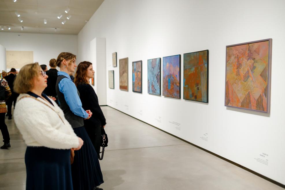 Visitors stand in a gallery space attentively viewing a row of abstract paintings displayed along a white wall; three women in the foreground, with other viewers continuing along the exhibition in the background.