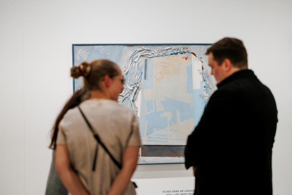 Divi apmeklētāji stāv galerijas zālē un no neliela attāluma aplūko gaišos toņos veidotu abstraktu gleznu ar fakturētu virsmu, kas eksponēta uz baltas sienas.