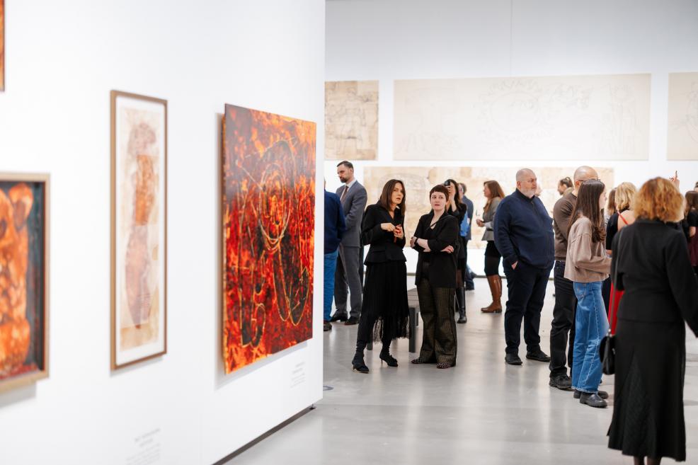 Galerijas zālē apmeklētāji pulcējas pie izstādes sienām, aplūkojot gleznas; priekšplānā redzama koši sarkanos un oranžos toņos veidota abstrakta glezna, fonā cilvēki sarunājas un vēro darbus.