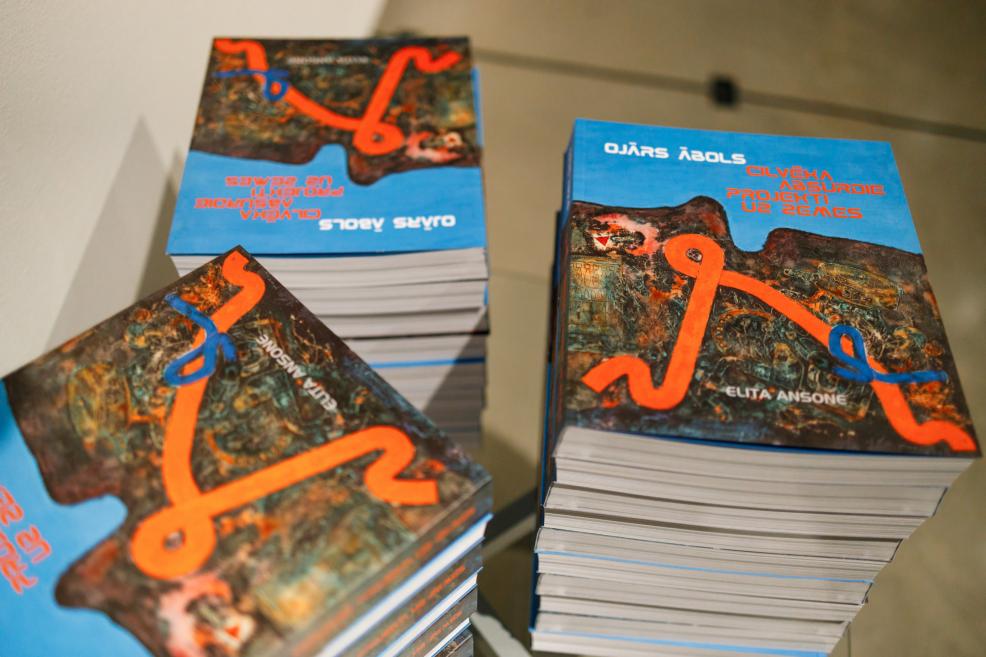 Uz galda sakrautas vairākas izstādes kataloga grāmatas ar nosaukumu “Ojārs Ābols. Cilvēka absurdi projekti uz zemes”, ar spilgtu zilu vāku un oranžu abstraktu līniju motīvu.