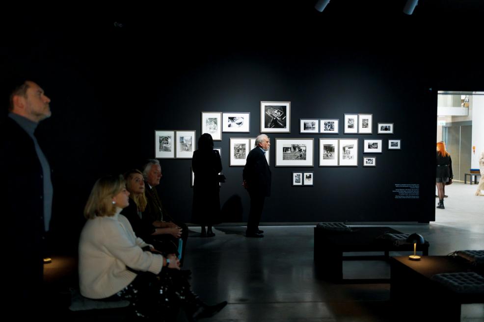 A dark exhibition gallery with a black wall displaying a composition of black-and-white photographs; visitors sit and stand in the foreground, quietly observing the works.