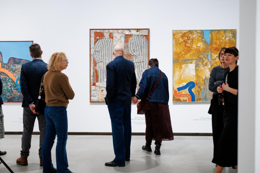 Exhibition visitors in a gallery space viewing paintings by Ojārs Ābols displayed on a white wall, with people standing and discussing the artworks in front of them.