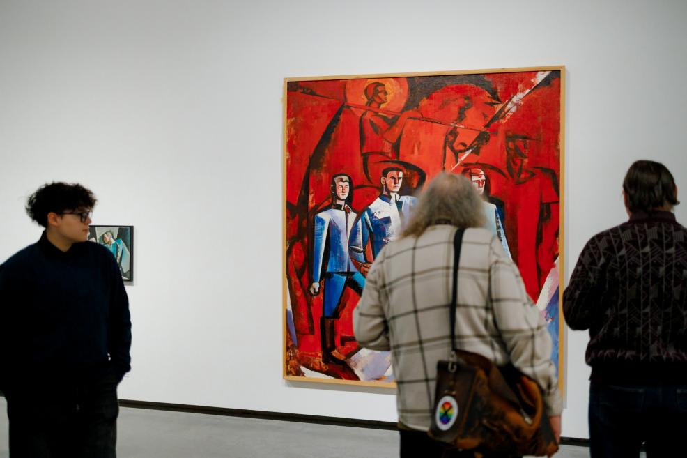 Izstādes apmeklētāji galerijas telpā aplūko lielizmēra gleznu ar dominējošu sarkanu krāsu un stilizētām cilvēku figūrām Ojāra Ābola daiļradē, kas eksponēta uz baltas sienas.