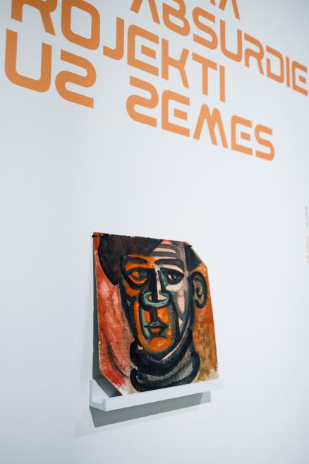 Uz baltas izstāžu zāles sienas redzams Ojāra Ābola gleznojums ar stilizētu cilvēka seju oranžos, melnos un brūnos toņos, novietots uz neliela sienas plaukta, virs kura eksponēts izstādes nosaukums “Cilvēka absurdie projekti uz zemes”.