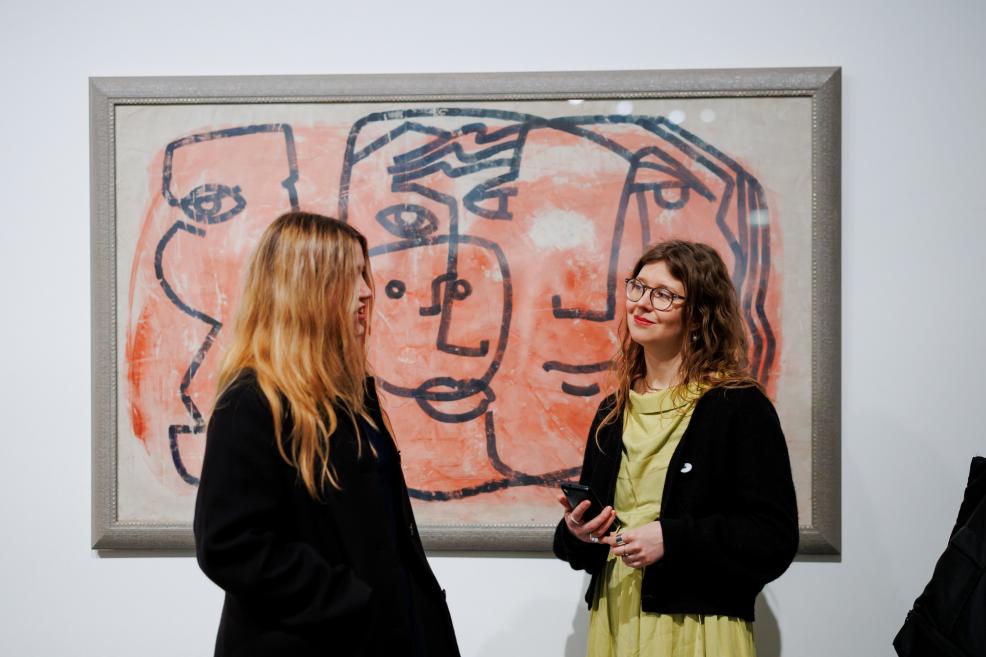 Two women are talking in an exhibition space while standing in front of a painting by Ojārs Ābols depicting stylised, overlapping human faces in pink and black tones, framed in grey.