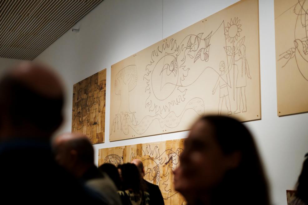 Large light-coloured drawings on paper with symbolic motifs—sun, plants, birds, and childlike figures—are displayed on an exhibition wall, with blurred silhouettes of visitors in the foreground.
