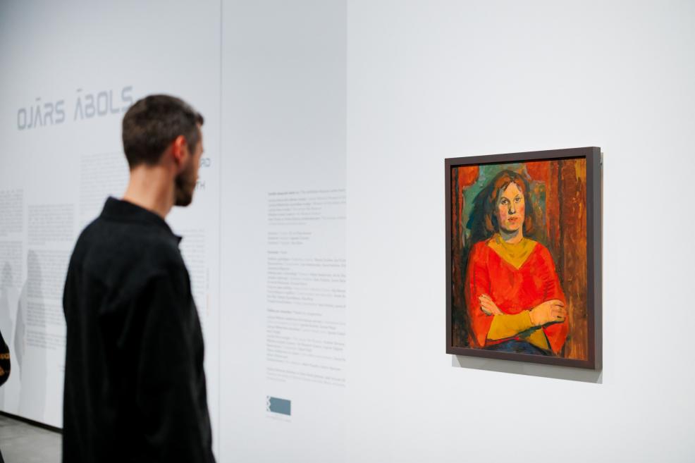 Apmeklētājs stāv izstāžu zālē pie sienas un aplūko nelielu gleznu ar sievietes portretu sarkanā apģērbā, fonā redzami izstādes teksti uz baltas sienas.