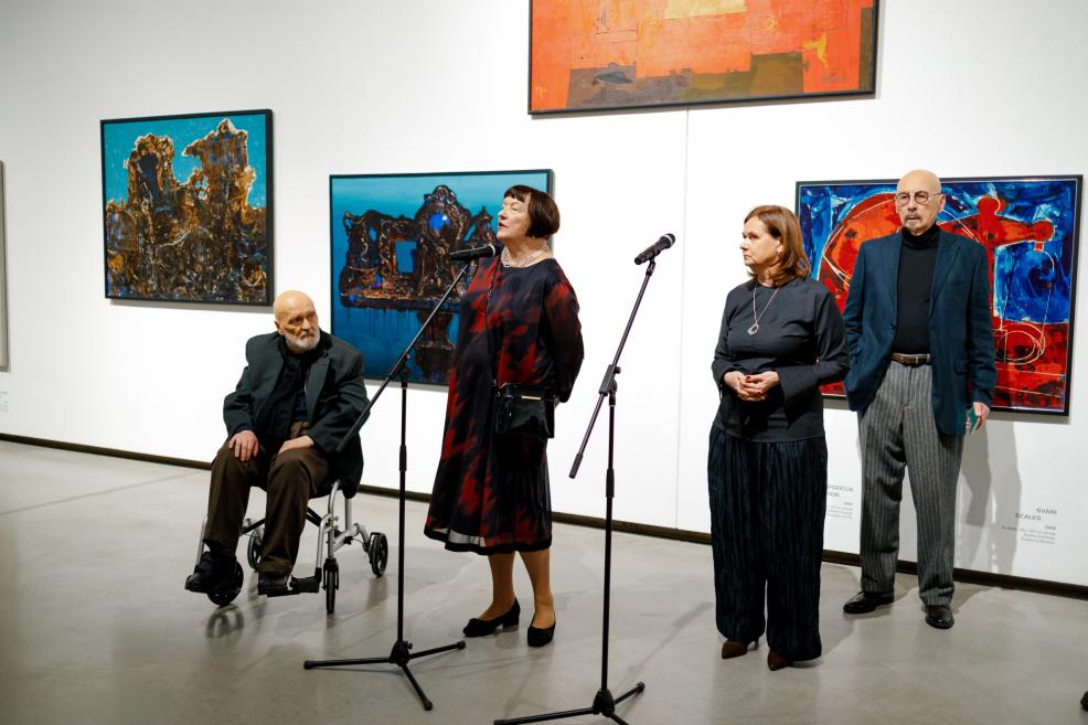 Izstāžu zālē četri cilvēki piedalās atklāšanas pasākumā: sieviete stāv pie mikrofona un uzrunā klātesošos, blakus atrodas vēl divi cilvēki, bet viens vīrietis sēž ratiņkrēslā; fonā redzamas krāsainas abstraktas gleznas uz baltas sienas.