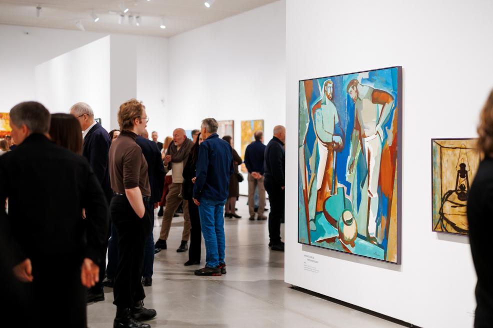 Visitors gather and converse in a spacious exhibition hall; on the right, a colorful figurative painting is displayed on a white wall, with additional artworks visible in the background.